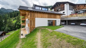 Een modern huis met een balkon, een garage met auto's en een bergachtig landschap op de achtergrond.