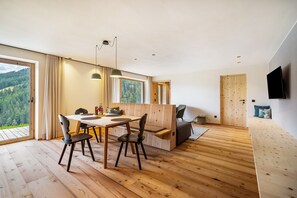 Moderni huone, jossa on puulattia, ruokapöytä tuoleineen, sohva, televisio ja suuri ikkuna, josta avautuu näkymä metsään.