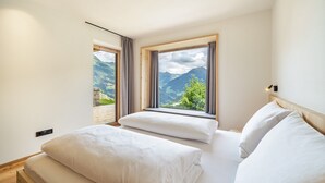 Een slaapkamer met een bed, twee ramen met gordijnen, uitzicht op bergen en een houten hoofdeinde.