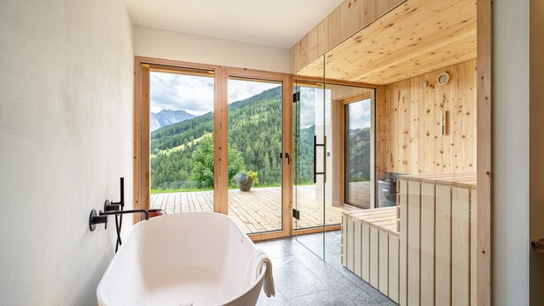 Een moderne badkamer met een vrijstaand bad, houten wandbekleding en een grote schuifdeur naar een terras met uitzicht op een berg.
