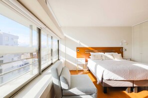 A modern hotel room with a large window, a bed, a bedside table, and a small seating area. - Elite Studio c/Smart TV & Wifi (Las Condes)