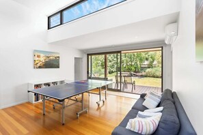A modern room with a ping pong table, a sofa, and a large window offering a view of the outdoors. - 5BR Modern Beach House Oasis (Cowes)
