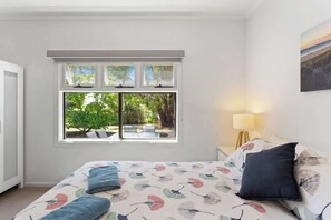 A bedroom with a bed, a window with a view of greenery, a bedside lamp, and a painting on the wall.