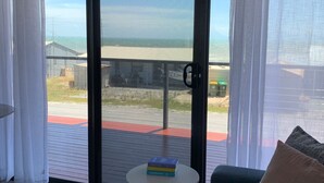 Ein Zimmer mit einer Schiebetür zu einem Balkon mit Blick auf Strand und Meer. Ein kleiner runder Tisch mit einem Buch darauf, eine Couch mit Kissen und ein gemustertes Teppich sind vorhanden.