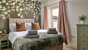 A bedroom with a bed, pillows, a nightstand, a dresser, a window with curtains, and a wall with a leaf pattern.