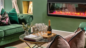 A green sofa with cushions, a glass table with a bottle and glasses, and a fireplace with a fire.