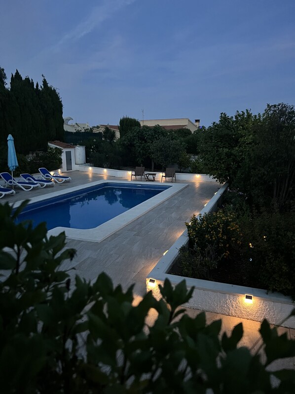A rectangular swimming pool with a tiled deck, surrounded by lounge chairs and a small table.