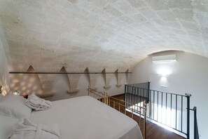 A bedroom with a large bed, a wooden floor, a balcony with a metal railing, and a stone ceiling.