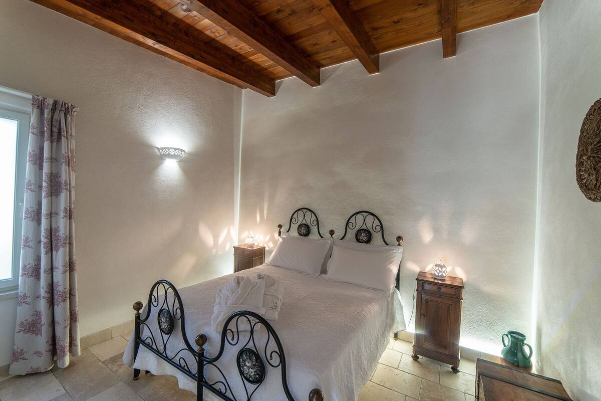 Un dormitorio con cama, cabecera de madera, mesitas de noche y una ventana con cortinas.