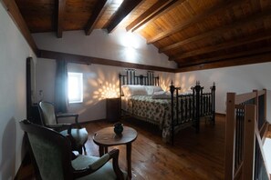 A bedroom with a wooden ceiling, a bed with a metal frame, a chair, a small table, and a window allowing natural light.