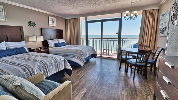 A hotel room with two beds, a dining table with chairs, a balcony with a view of the sea, and a chandelier.