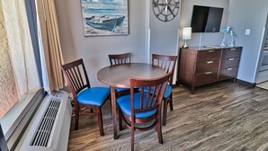 A dining area with a round table, four chairs with blue cushions, a wooden cabinet with drawers, a wall-mounted TV, and a nautical-themed painting.