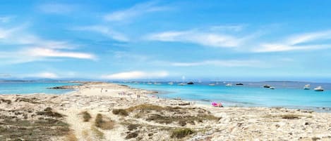 Una playa con aguas azules cristalinas, embarcaciones y vegetación escasa.