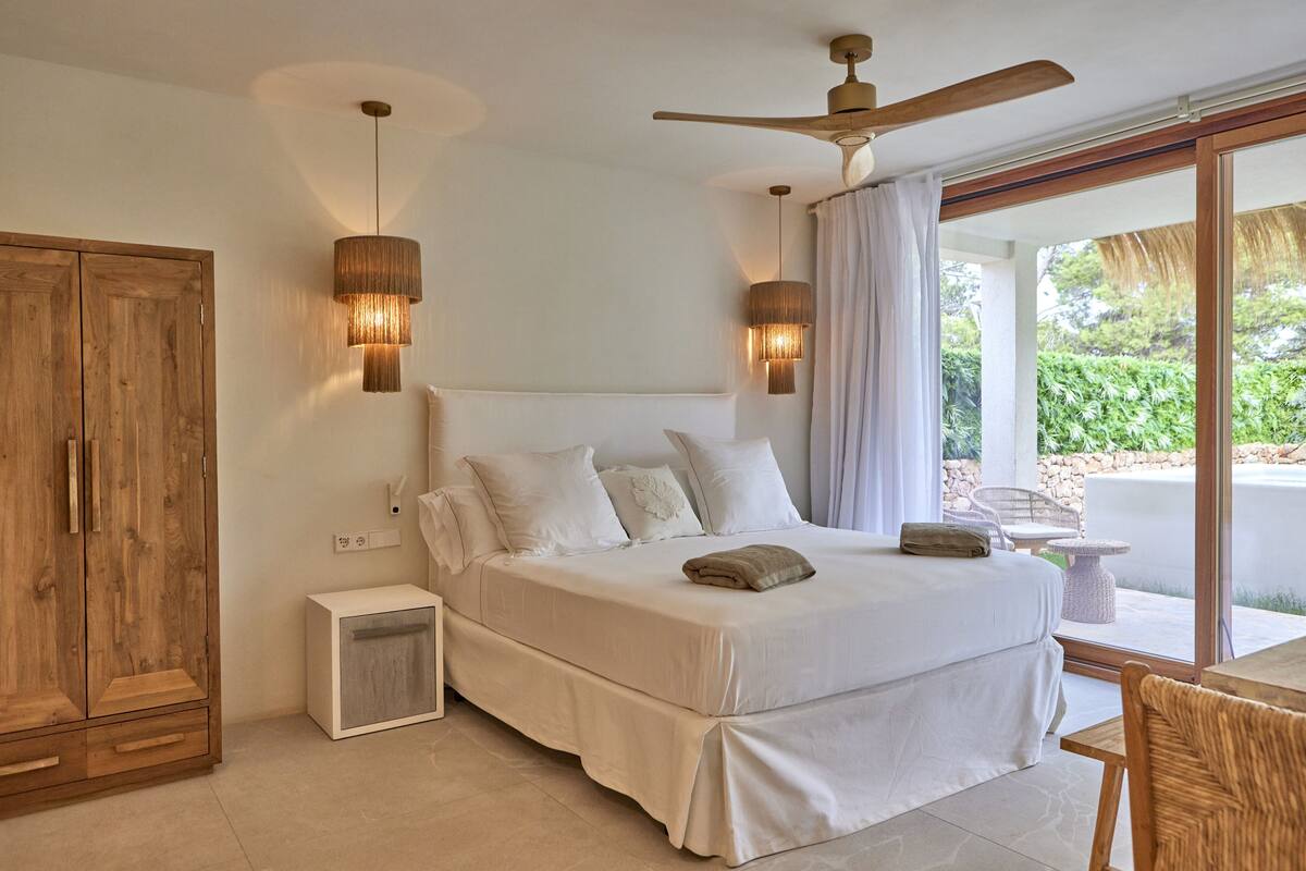 Quarto com cama grande, guarda-roupa de madeira, mesa de cabeceira e ventoinha no teto.