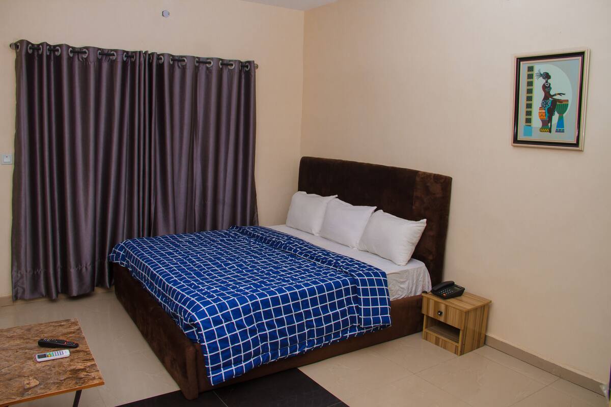 A bedroom with a bed, a nightstand, a remote control, and a curtain.