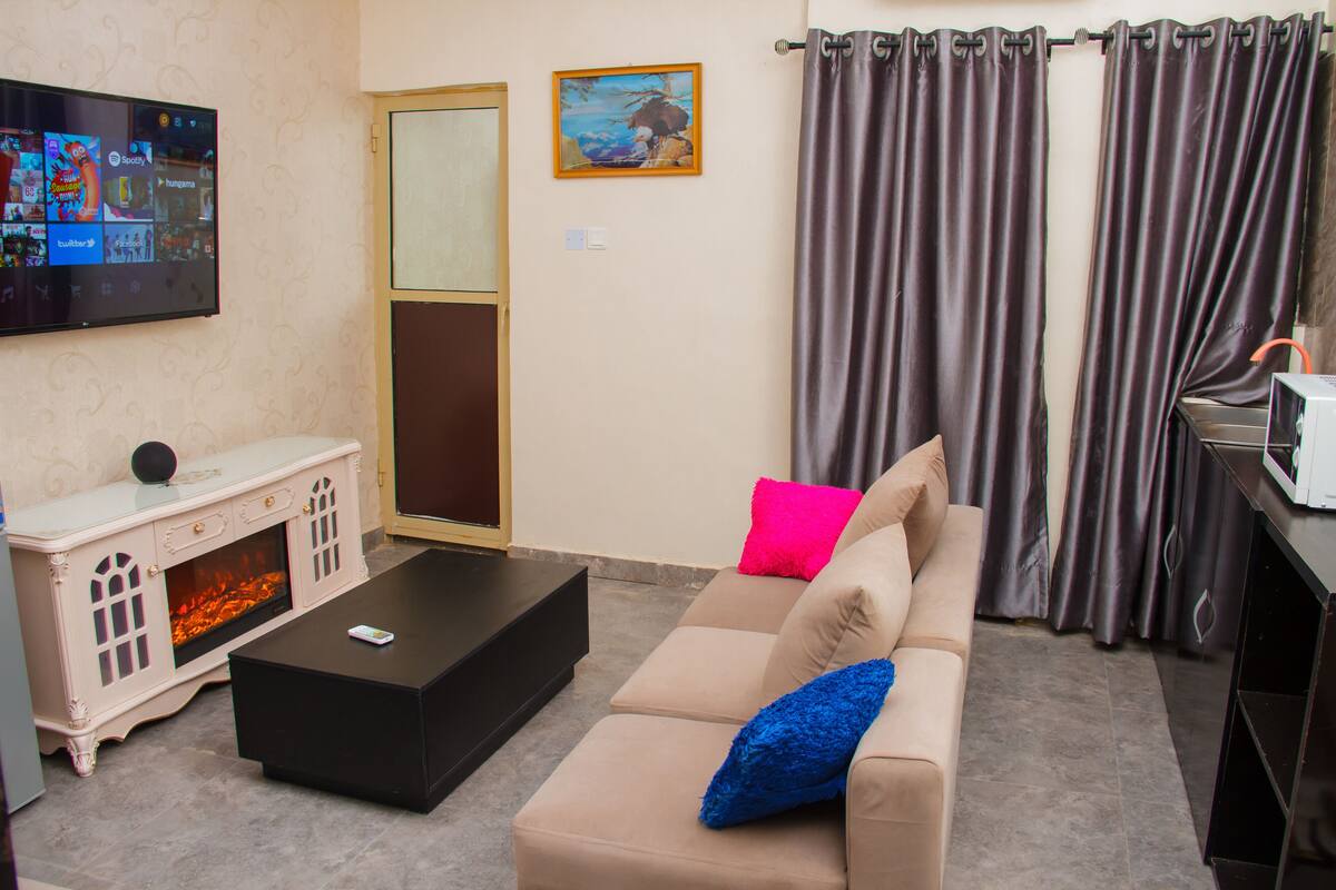 A living room with a flat-screen TV, a fireplace, a sofa with colorful cushions, and a curtain.