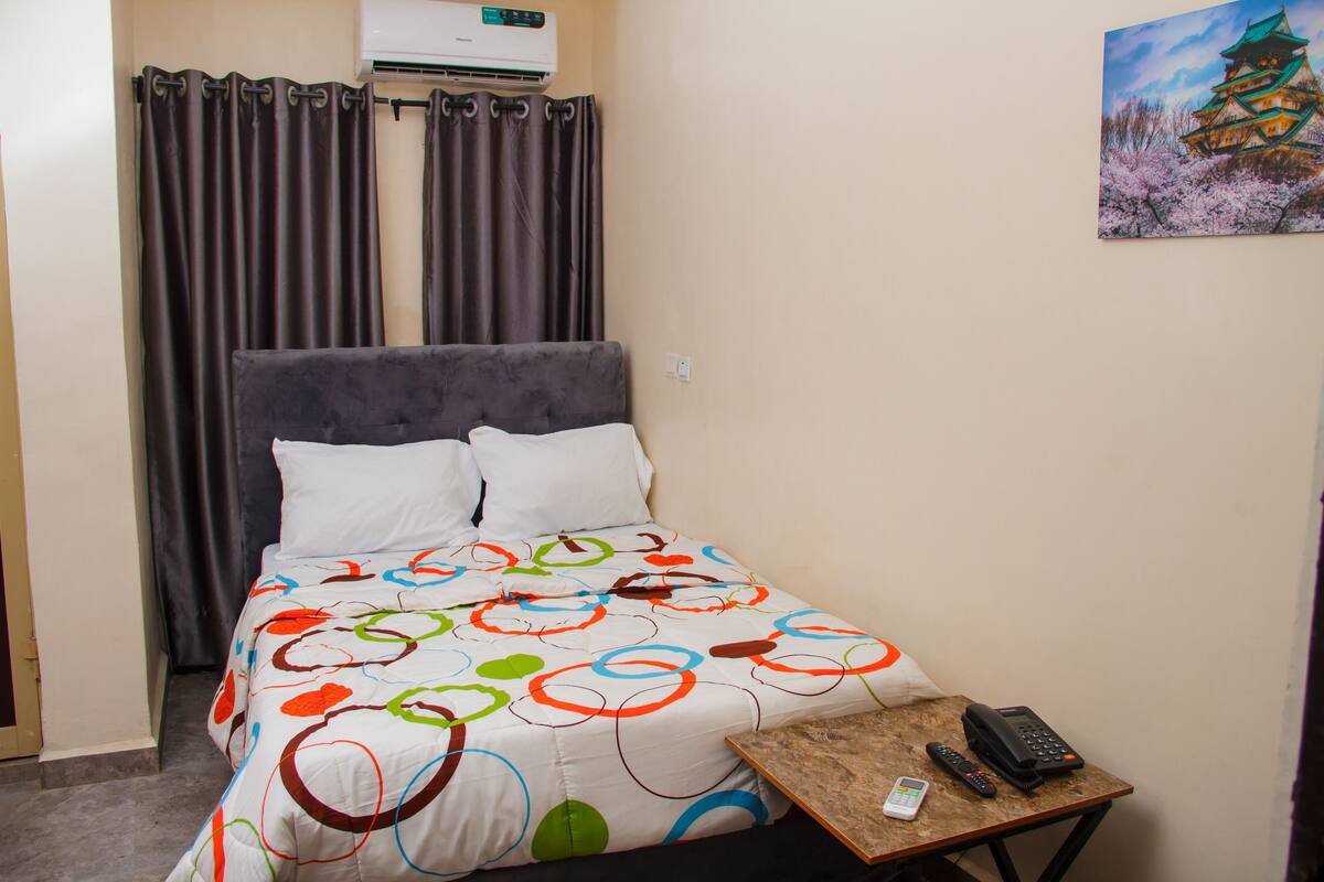 A hotel room with a bed, a small table with a phone and remote controls, and a painting of a traditional building on the wall.