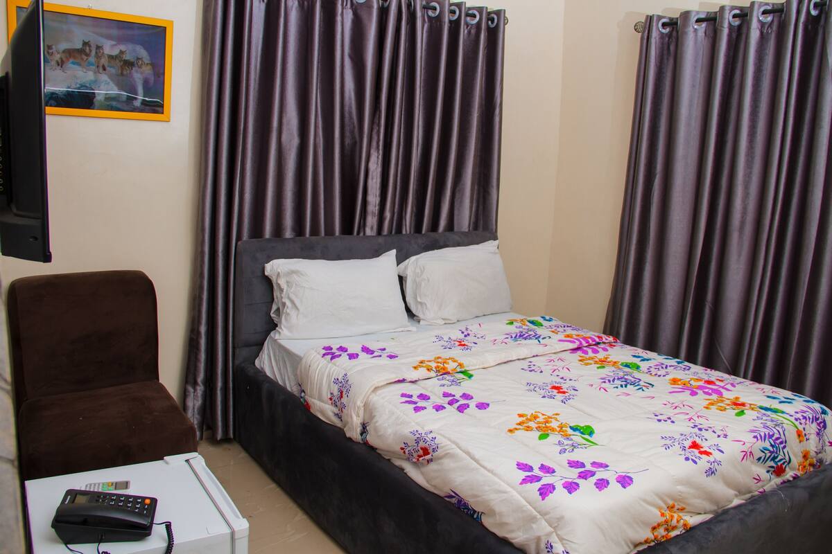 A bedroom with a bed, a chair, a small table with a phone, and curtains.