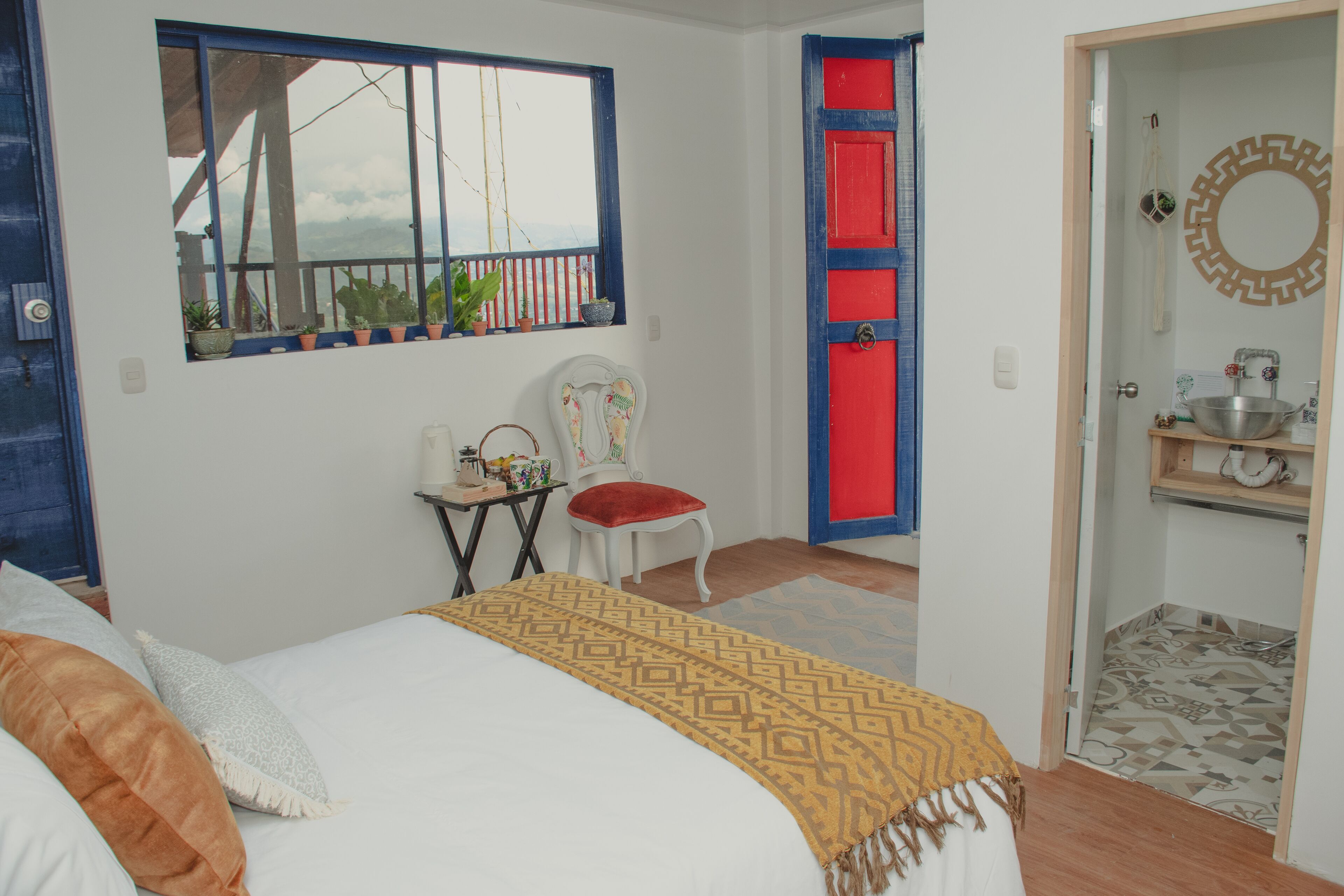 A bedroom with a bed, a chair, a small table, a window with plants, and a red door.