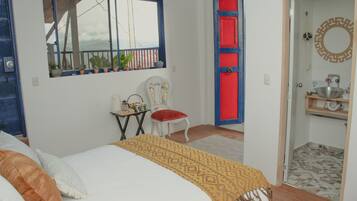 A bedroom with a bed, a chair, a small table, a window with plants, and a red door.