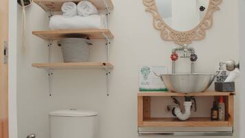 A bathroom with a toilet, a sink with a decorative mirror, and shelves with towels and toiletries.