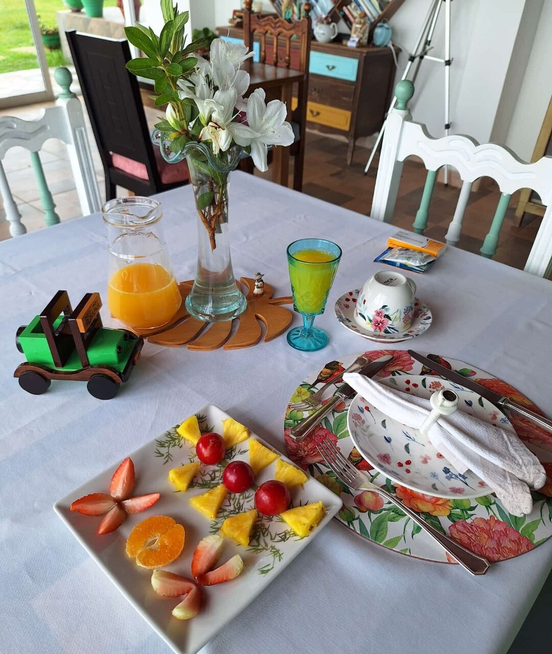 A breakfast table with fruit, a vase with flowers, a glass of orange juice, and a toy car.