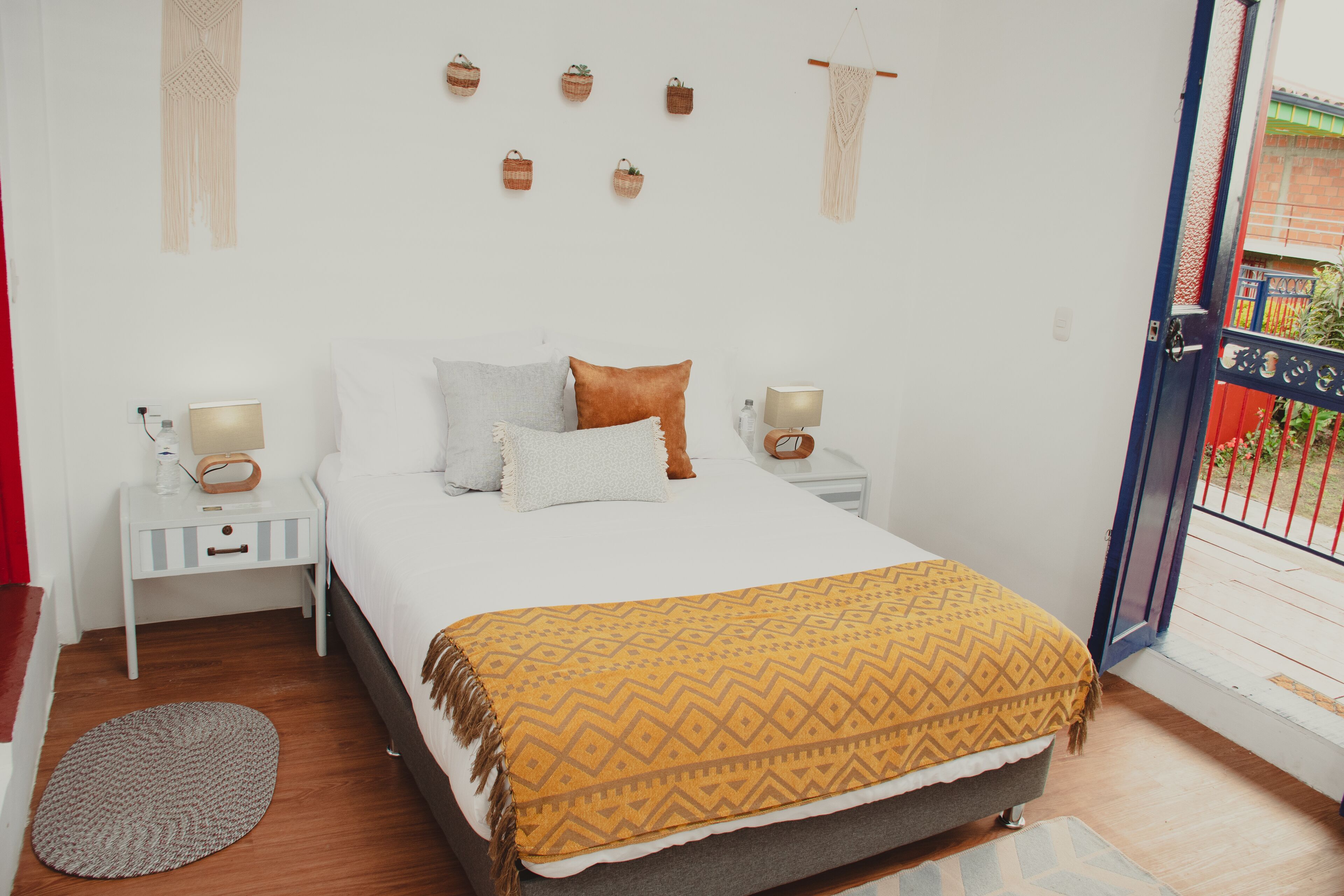 A bedroom with a bed, bedside tables, a bedside lamp, a patterned throw blanket, and a macrame wall hanging.