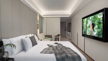 Une chambre d’hôtel moderne avec un grand lit, un téléviseur à écran plat fixé au mur, une petite table avec une plante et une vue sur la verdure environnante.