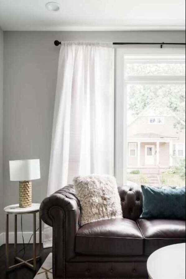 A room with a leather sofa, a small side table with a lamp, a window with a view of a house, and a curtain. - Luxurious 3,000 Square Foot House Downtown (Indianapolis)
