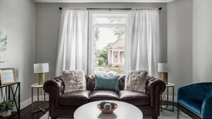 Un salon avec un canapé en cuir marron, une table basse ronde et une vue sur une maison, visible à travers une fenêtre ornée de rideaux blancs.