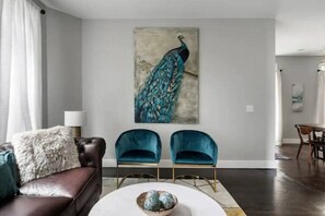 Uma sala de estar com sofá de couro marrom, duas poltronas de veludo azul, uma mesa de centro branca e um quadro com a imagem de um pavão na parede.