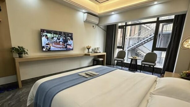 Une chambre d’hôtel avec un grand lit, un téléviseur fixé au mur, une petite table avec une chaise et une fenêtre donnant sur un immeuble.