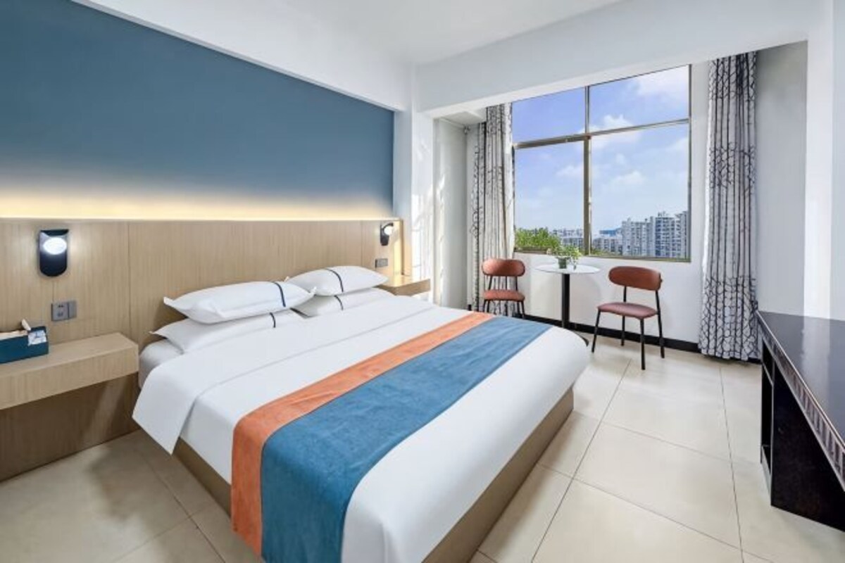 A hotel room with a bed, bedside table, chair, desk, and a large window with a city view.