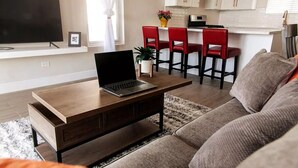 Un salon avec une table basse en bois, un ordinateur portable, un canapé et un téléviseur. On aperçoit une cuisine à l’arrière-plan, avec des chaises rouges et une plante.