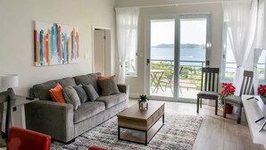 Un salon moderne avec un canapé gris, des fauteuils rouges, une table basse et une vue sur la mer grâce à de grandes portes vitrées.
