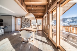 A spacious wooden interior with a dining area, stone wall, and large windows offering a view of a snowy landscape.