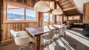 A wooden interior with a dining table, chairs, and a sofa. Large windows offer a view of a snowy landscape.