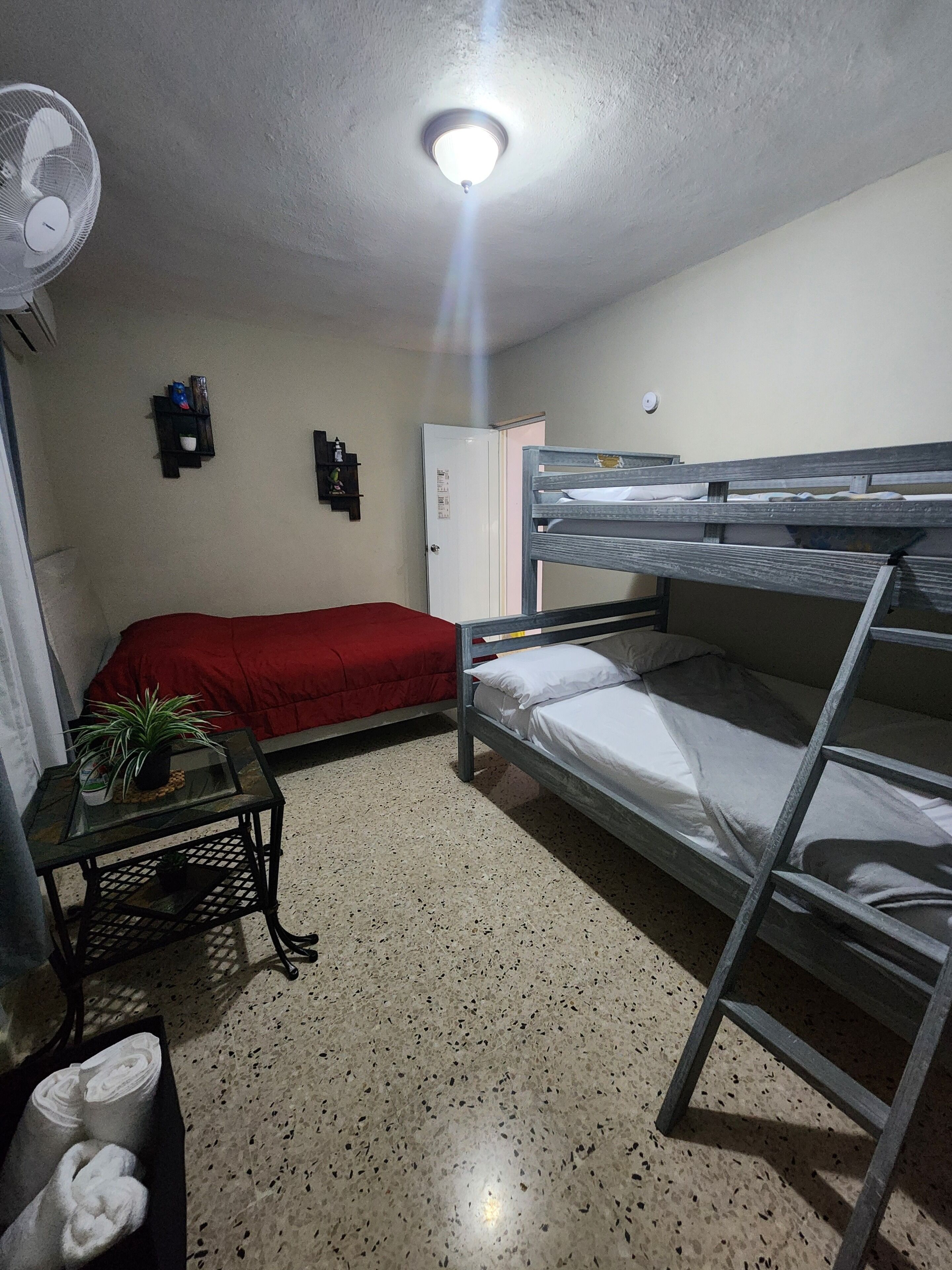 Un dormitorio con literas, un escritorio con una planta, un ventilador y una puerta.