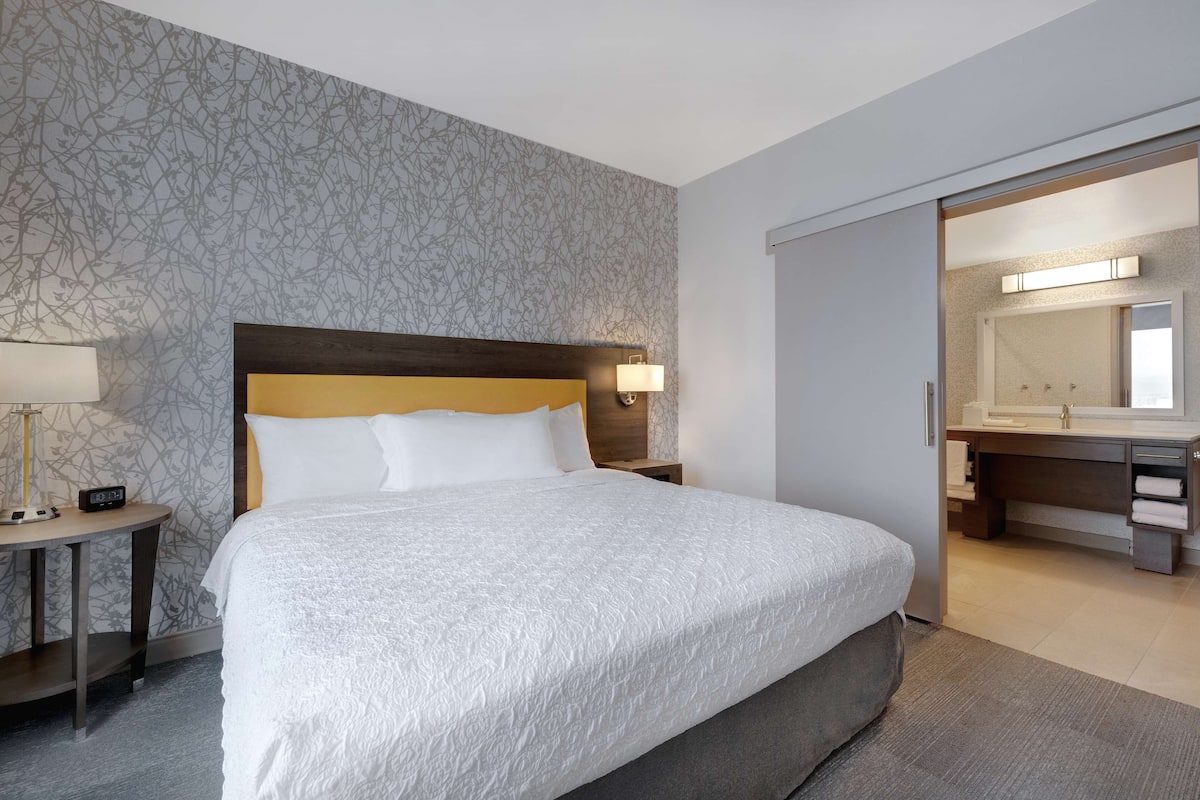 Ett hotellrum med en stor säng, ett sängbord med en lampa, ett litet bord och ett badrum som syns genom en öppen dörr.