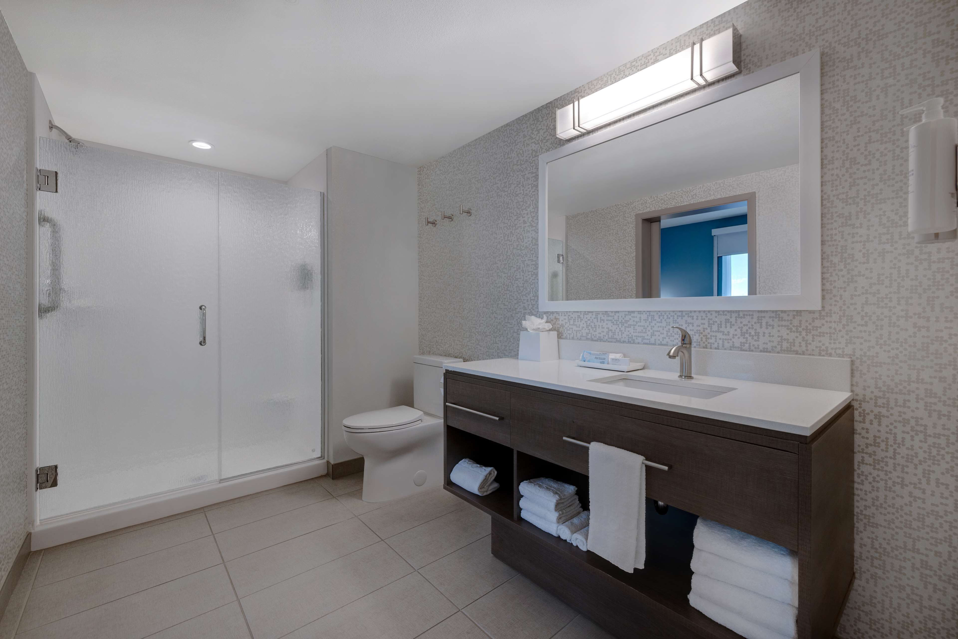 Une salle de bain moderne avec une douche à l’italienne, des toilettes, un meuble-lavabo avec un grand miroir et des serviettes soigneusement pliées.