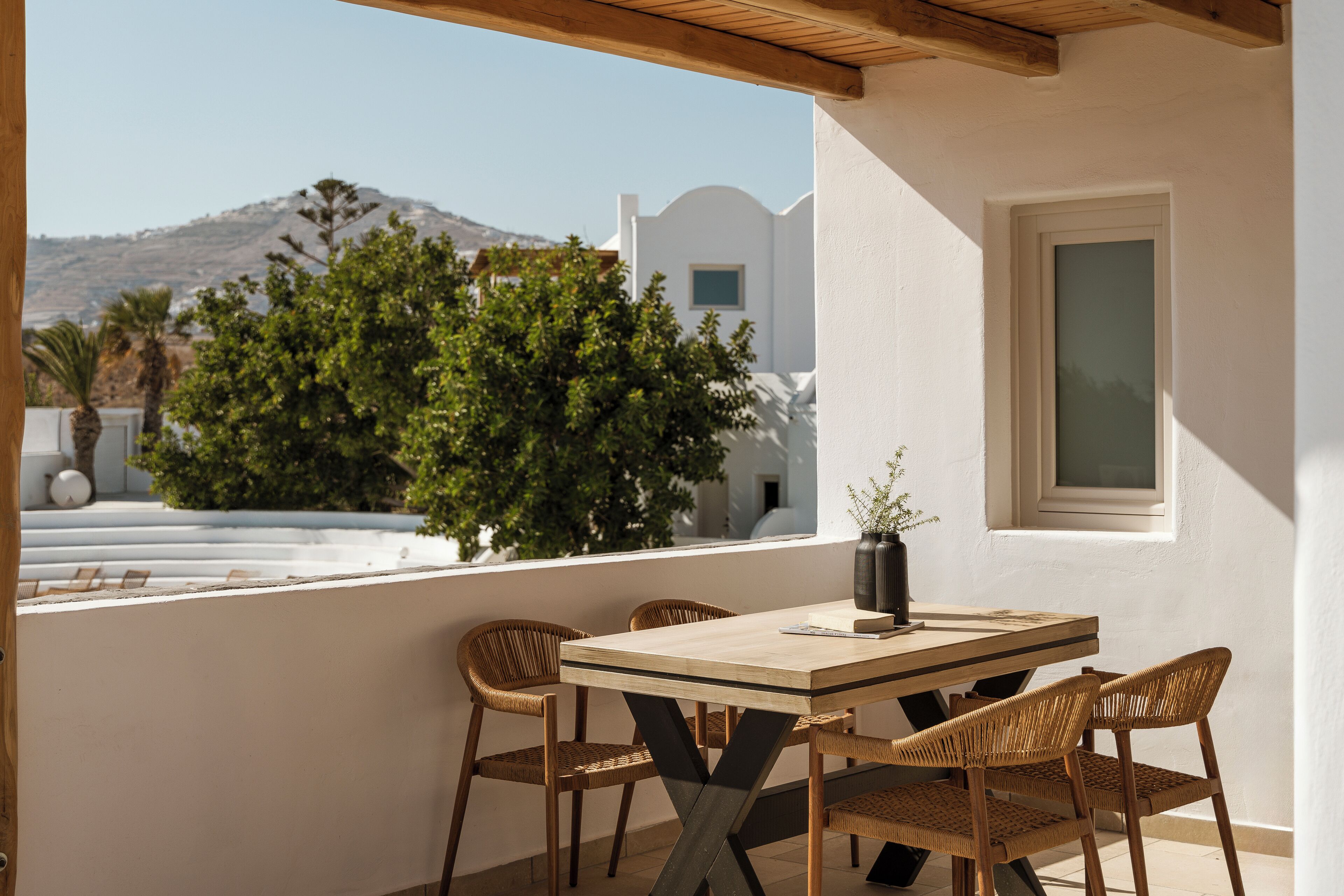 A balcony with a wooden table and wicker chairs, overlooking a white building and a tree.
