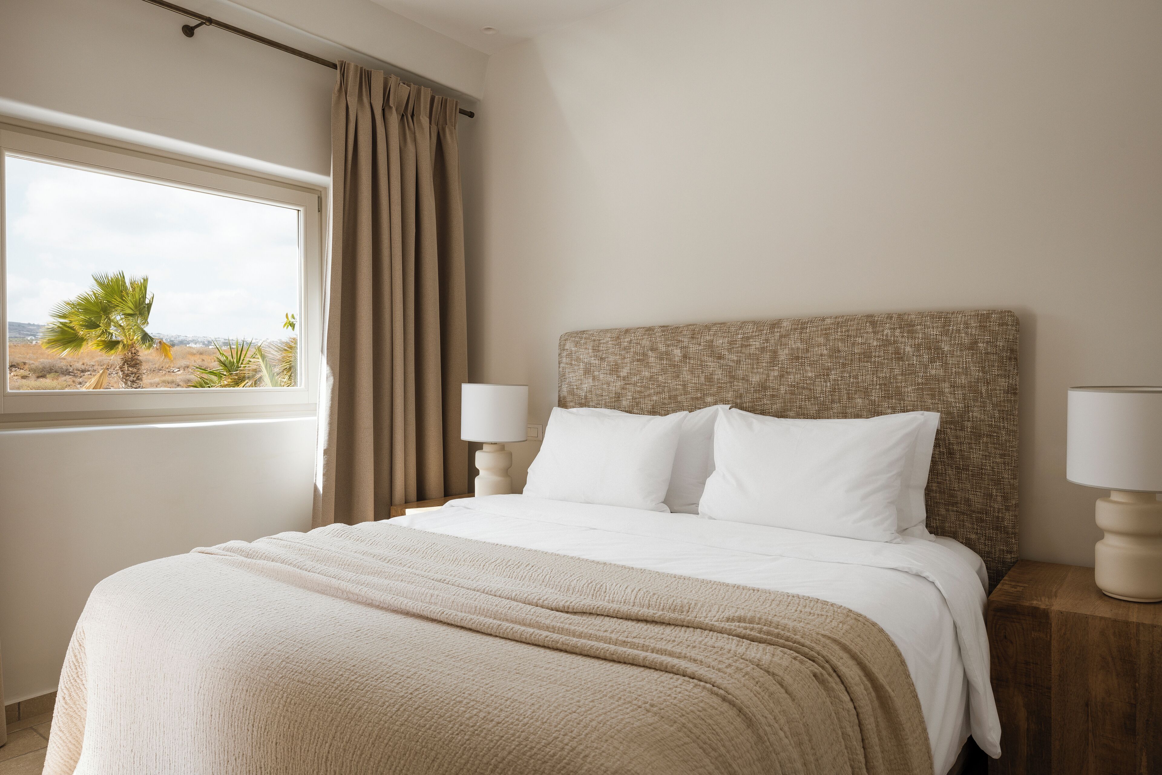 A neatly made bed with a beige blanket, a headboard with a textured pattern, two bedside lamps, and a window with a view of a palm tree and distant landscape.