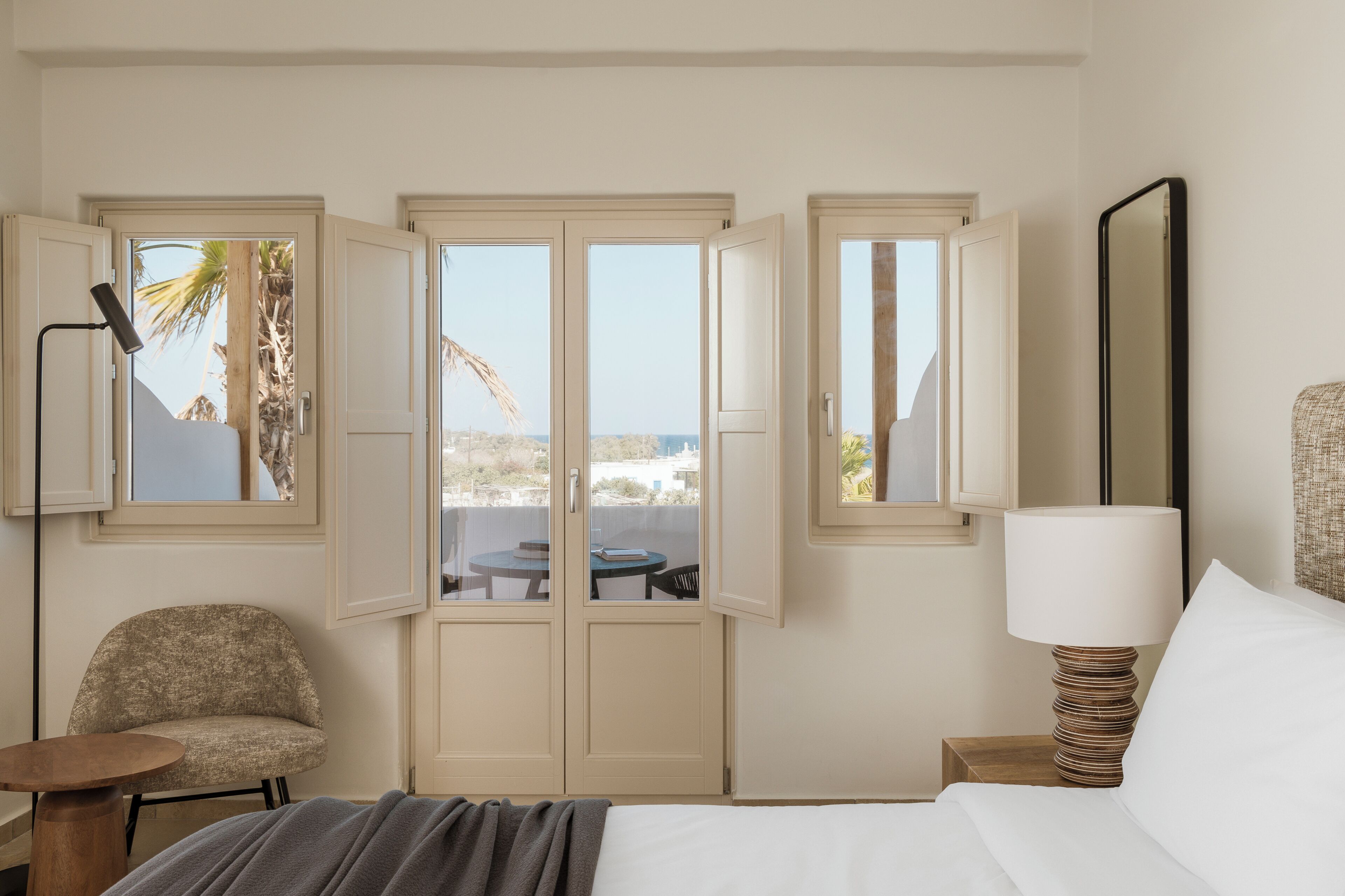 A hotel room with a bed, a chair, a table, a lamp, and a view of palm trees and buildings through open doors.