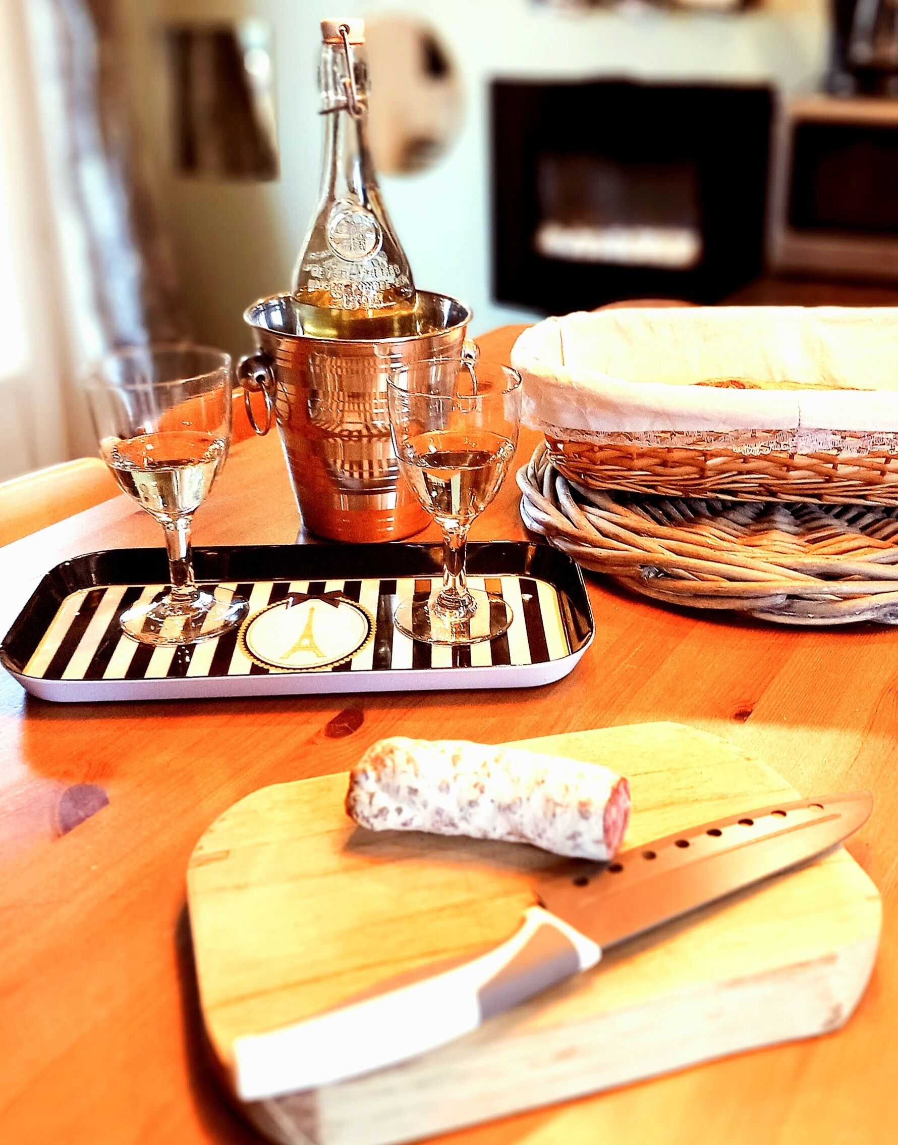 A wooden table with a bottle, two glasses, a tray, a basket, and a knife.