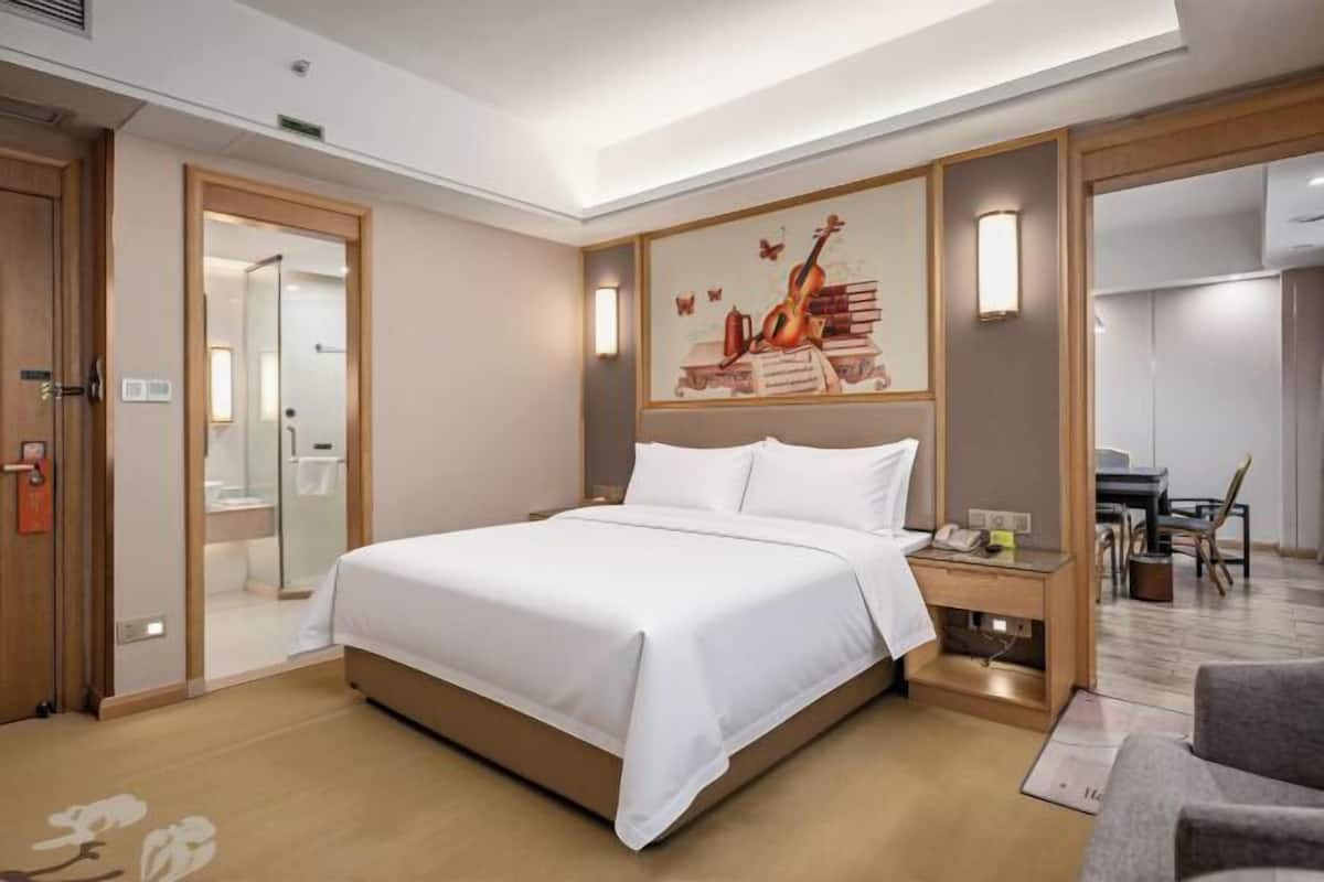 A hotel room with a large bed, a desk with a chair, a painting on the wall, and a bathroom visible through an open door.