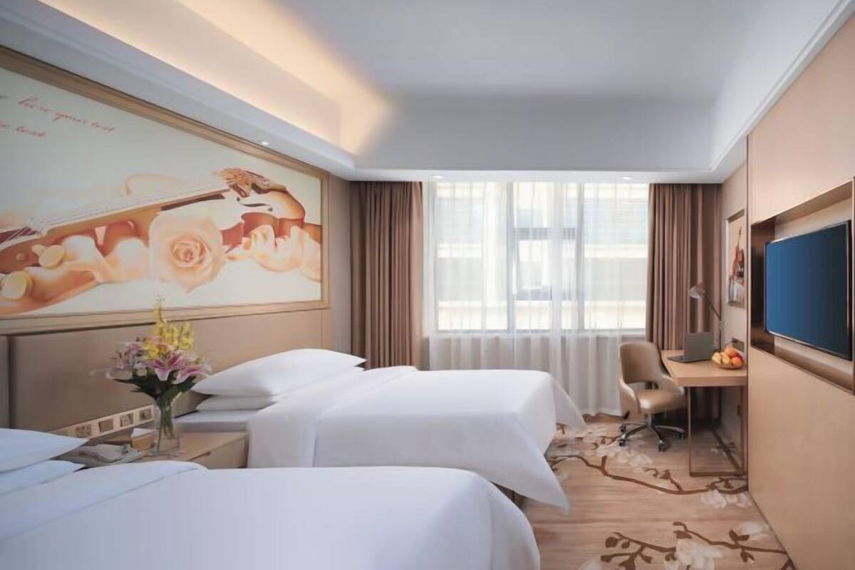 A hotel room with two beds, a desk with a laptop, a TV, and a large window with curtains.