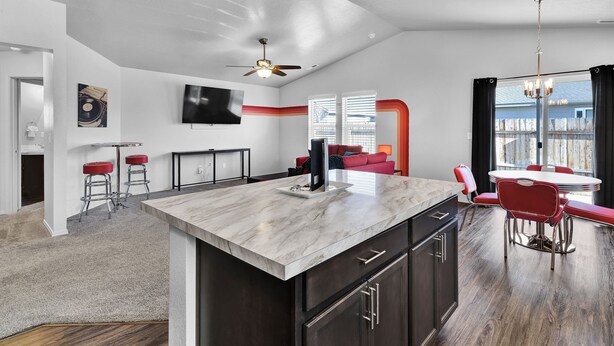 A modern kitchen with a marble island, dark cabinets, and a dining area with red chairs. - Monochrome Home Near Hospital (Nampa)