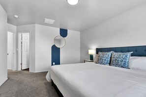 Une chambre d’hôtel avec un grand lit, une table de chevet avec une lampe, un mur d’accent bleu et une porte donnant sur une autre pièce.