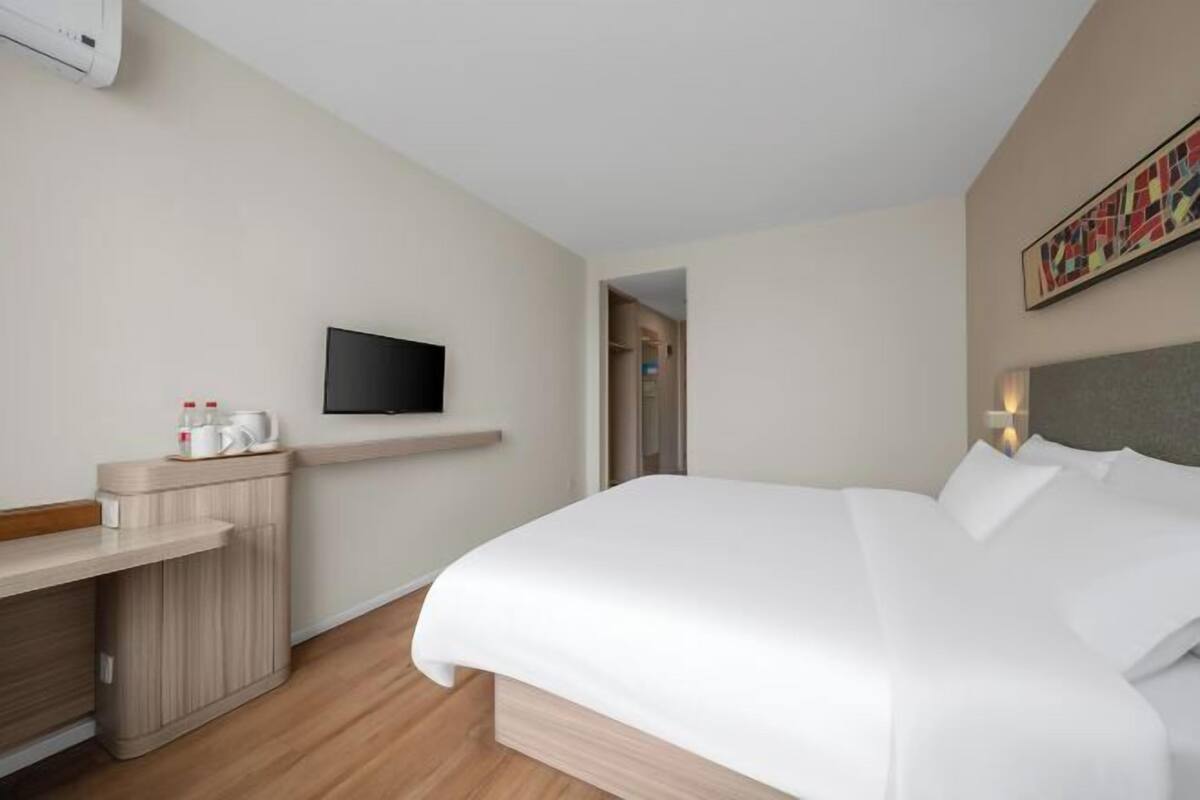 A hotel room with a bed, a TV mounted on the wall, a small desk with a coffee maker, and a painting on the wall.