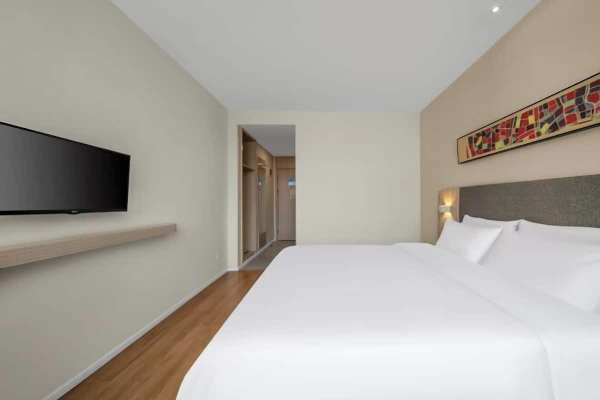 A hotel room with a bed, a mounted TV, a painting on the wall, and a hallway leading to another room.